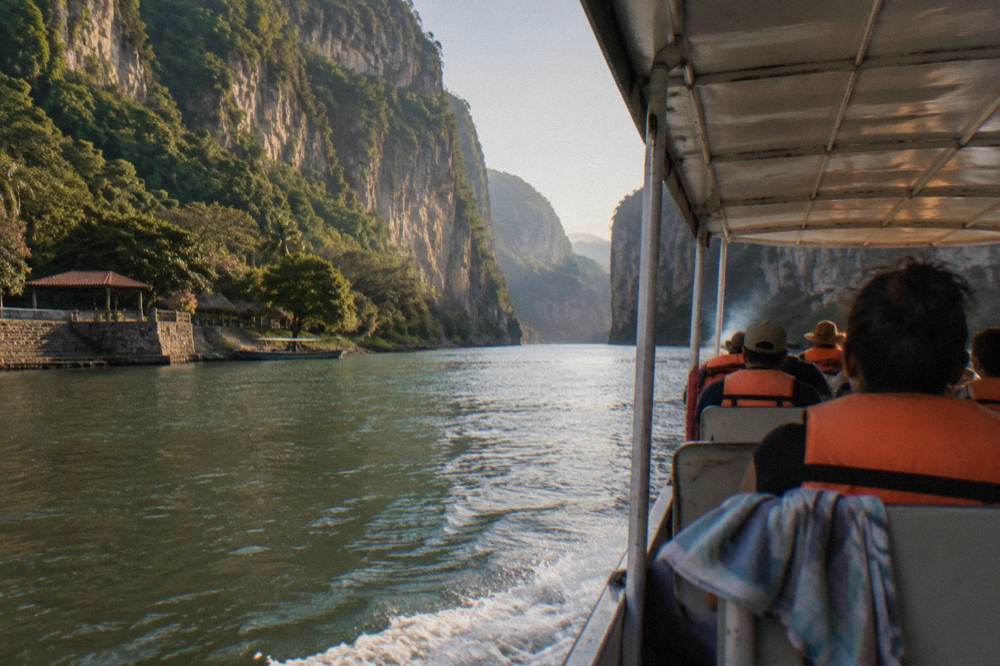 A covered motorboat carrying passengers along a wide river between towering canyon cliffs in warm morning light