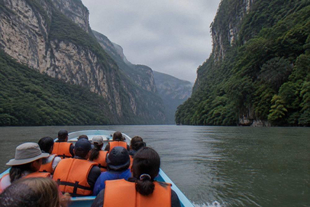 A long open boat carrying passengers along a wide river between steep forested canyon walls