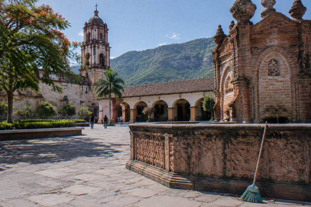 A colorful colonial town square with a brick fountain, arcades and a church tower under a bright sky