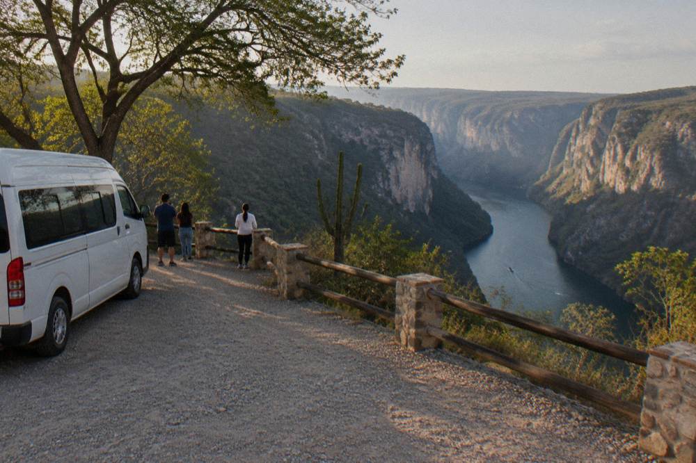 Air-conditioned tour van parked at a viewpoint overlook above the canyon with hills beyond