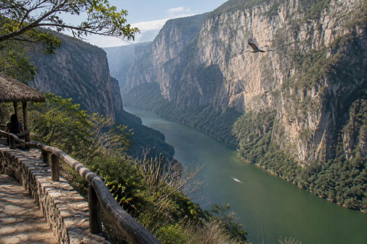 Tall limestone canyon walls rising above a wide green river with a small boat far below