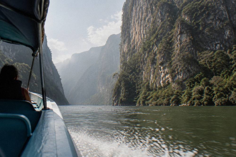 Speedboat on a wide river between towering forested canyon walls under hazy sky