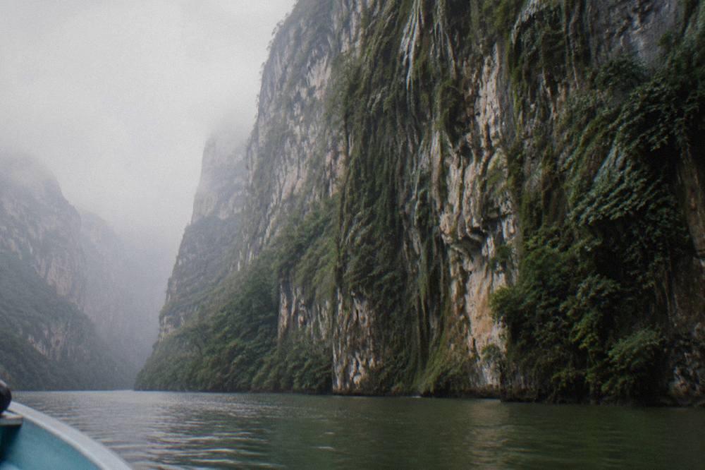 Sheer rock cliff face covered in green vegetation dropping to a calm river below