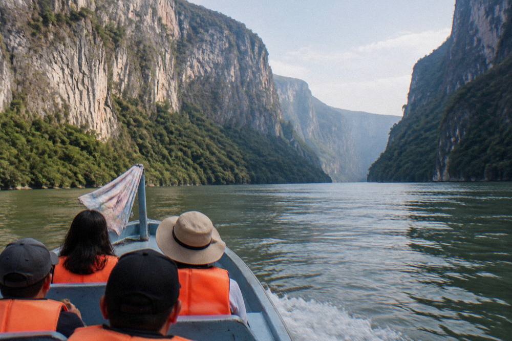 A small boat traveling on a wide river between towering canyon walls at midday