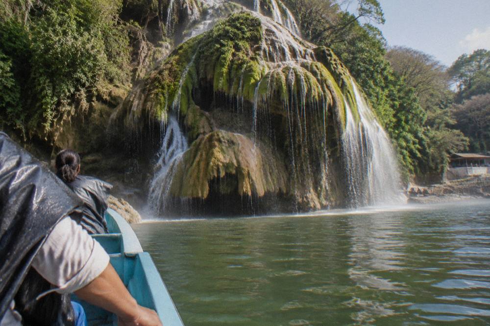 A flat-bottomed tour boat cruising close to a tall mossy cliff face above calm green water