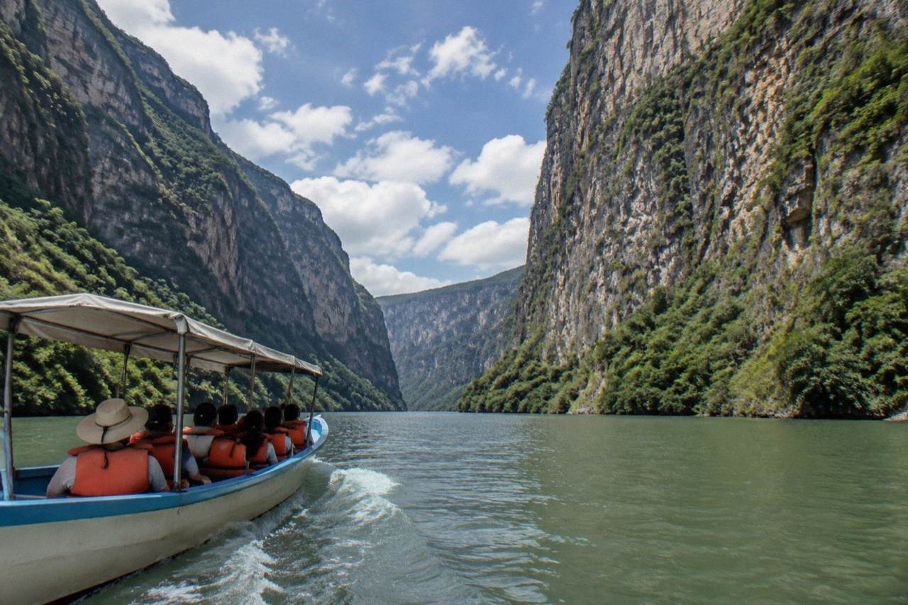 A boat on a wide river between tall limestone canyon walls under a bright sky with scattered clouds