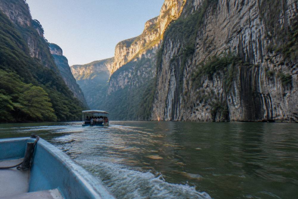 Covered tourist boat on a wide river heading toward tall cliff walls in morning light