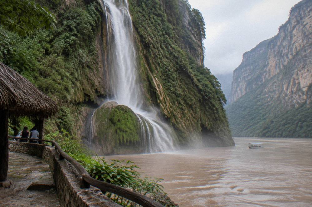 Tall waterfall falling from the top of a green canyon wall onto a wide brown river