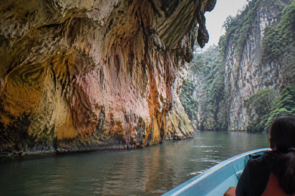 Small cave with mineral stained walls opening onto a calm river between cliffs
