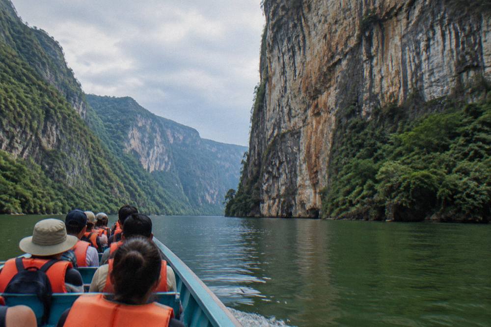 A long open tour boat cruising on calm river water beneath towering rock cliffs