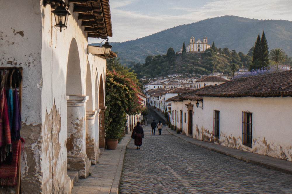 A colonial street with white arched facades and a hillside church in warm afternoon light