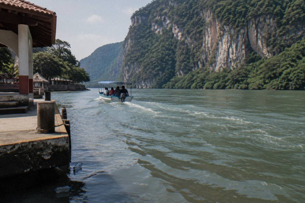 A small shared boat moving along a wide river between steep forested canyon cliffs