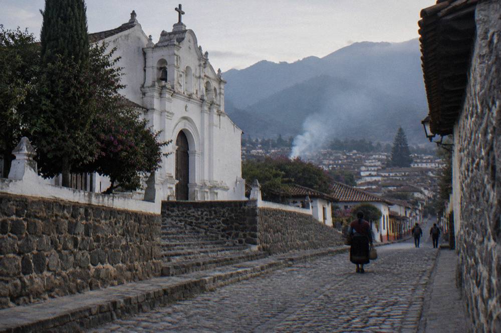 A highland village street with white church facade and mountains behind
