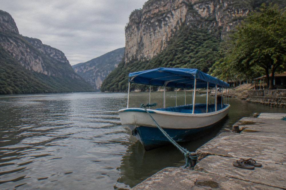 Blue and white covered passenger boat moored at a riverside dock with canyon cliffs behind
