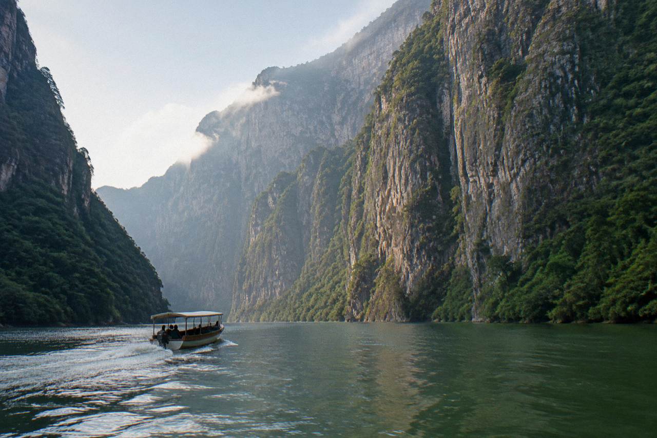 Wooden boat crossing a wide river between tall limestone canyon walls in morning light