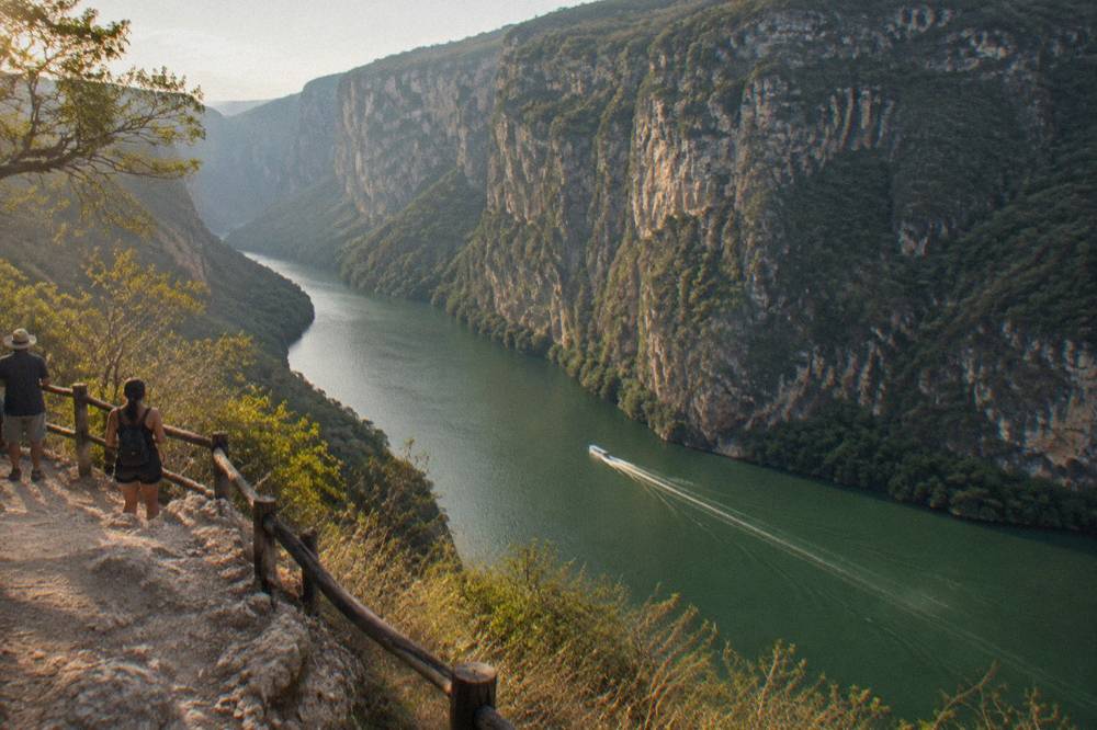 A long boat wake crossing a wide green river toward towering rock cliffs