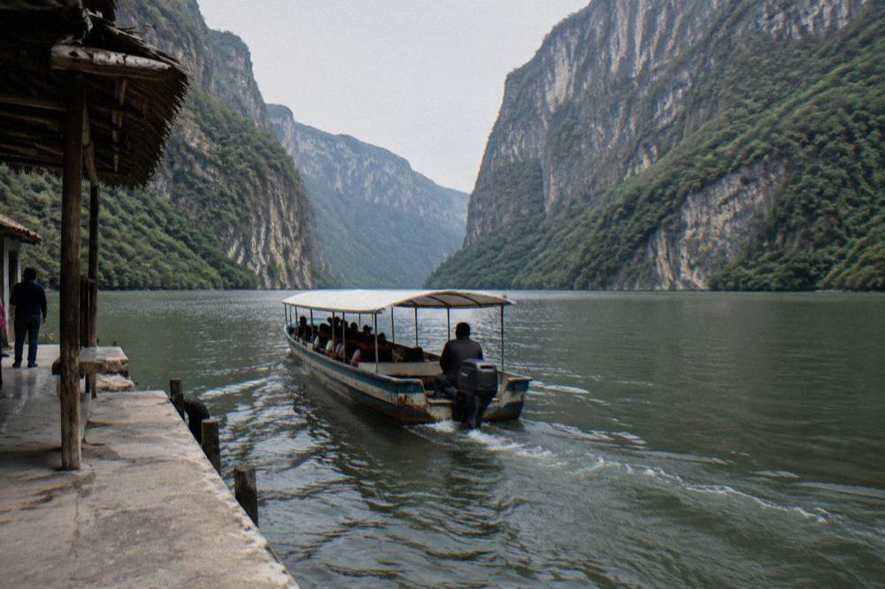 A long covered tour boat on a wide river between steep forested canyon walls