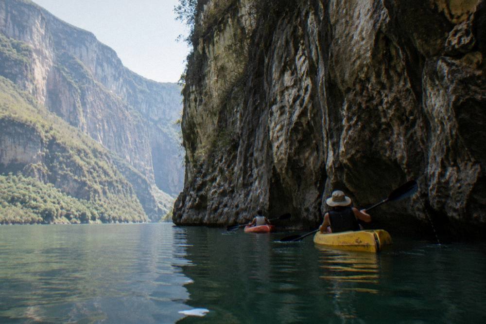 Two kayaks paddling close to a tall shadowed rock wall above still water